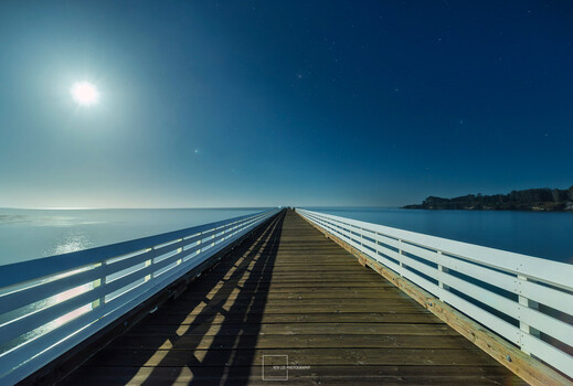 Long wooden pier with white railings extending toward the horizon under bright sunlight