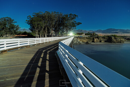 White wooden railings frame a curved bridge over still water with eucalyptus trees and mountains beyond