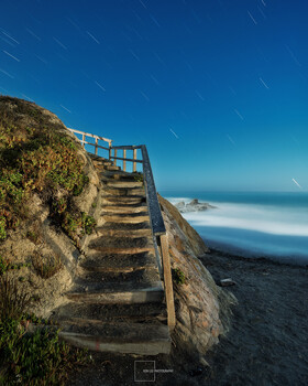 Wooden staircase descending a moss-covered cliff toward a misty coastal beach under clear blue sky