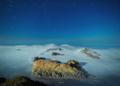 Rocky outcrops emerging from fog-covered valley at night with star trails above