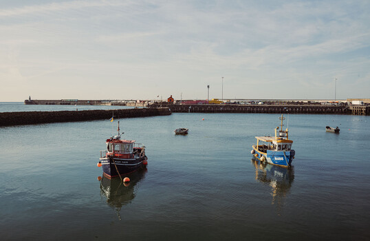 Two fishing boats moored in calm harbor water with industrial pier structures in background