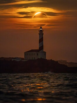 Lighthouse silhouetted against golden sunset with sun partially obscured by clouds above water
