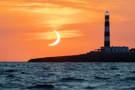 Crescent moon silhouetted against orange sunset sky above a striped lighthouse on a rocky coastal peninsula