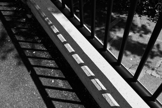 Strong shadow patterns cast across a white concrete ledge and pavement by metal railings and sunlight
