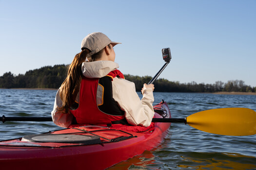 Woman in kayak taking selfie with paddle, wearing red life vest and cap on calm blue water