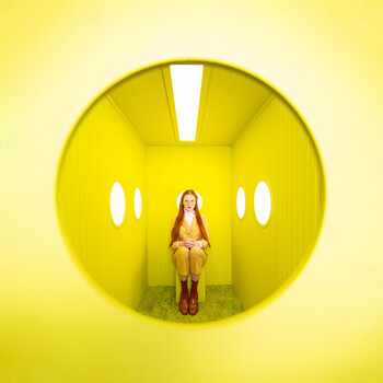 Woman seated inside a large yellow spherical tunnel structure with circular window openings