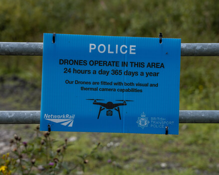Blue police drone warning sign mounted on metal rail with foliage background