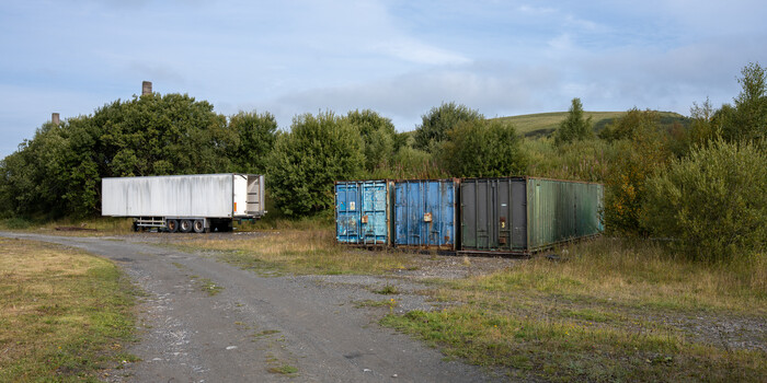 Abandoned shipping containers and a white semi-truck trailer parked on an overgrown lot with vegetation