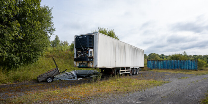 Abandoned Carrier refrigerated truck with lowered ramp on overgrown lot beside blue shipping containers