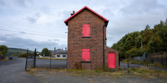 Abandoned brick tower with bright pink doors and shutters, surrounded by overgrown vegetation and industrial fencing