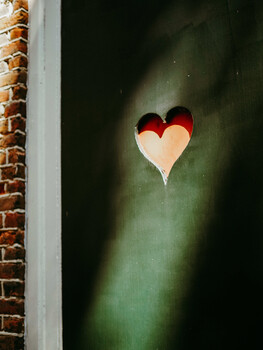Red and cream layered heart shape centered on textured green wall with brick visible at left edge