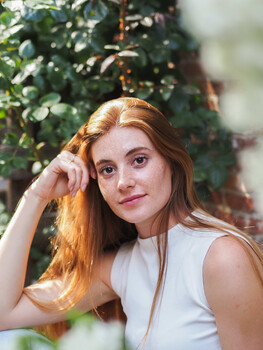 Woman with long copper hair posing against ivy-covered wall in natural light