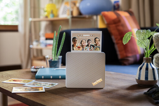 Instax instant camera displayed on wooden desk with photo prints and plants in home office setting