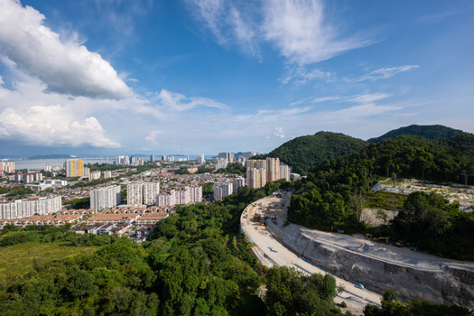 Aerial view of a curved elevated highway cutting through lush green forest with cityscape and mountains in the distance