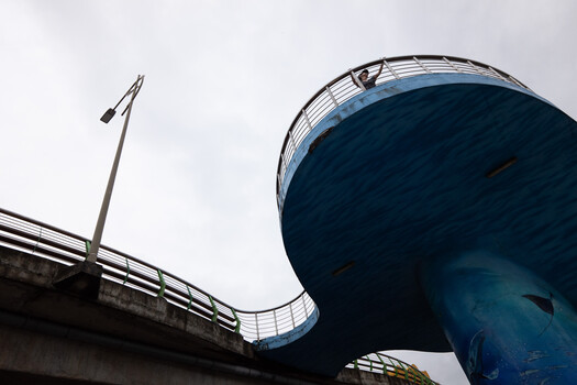 Blue water tank elevated on a concrete support structure with metal railing and flagpole against overcast sky