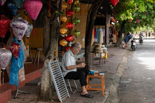 Elderly man sitting on wooden stool playing keyboard beside colorful lanterns on Vietnamese street