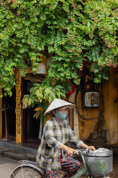 Woman in conical hat and plaid shirt sitting on bicycle with basket in front of ivy-covered building