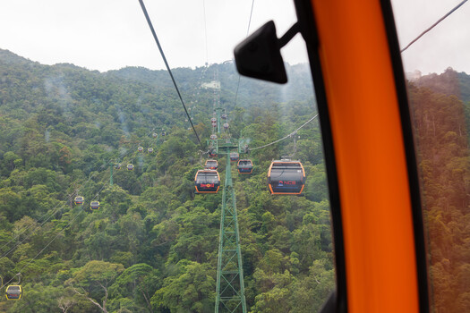 View from inside a cable car cabin showing suspended red gondolas crossing a forested mountain valley
