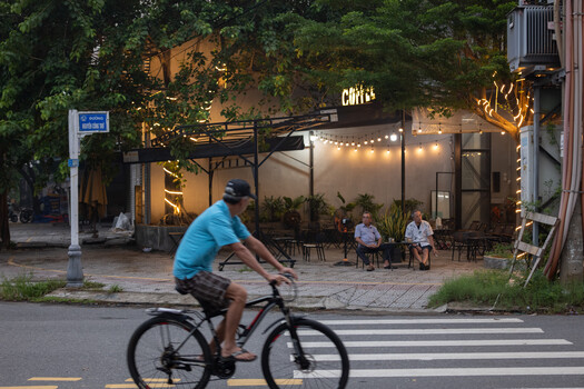 Cyclist in blue shirt riding across crosswalk in front of illuminated café storefront at dusk