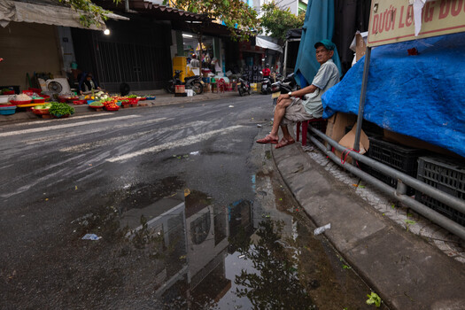 Man sitting on sidewalk next to blue truck in urban street market with produce and vendors
