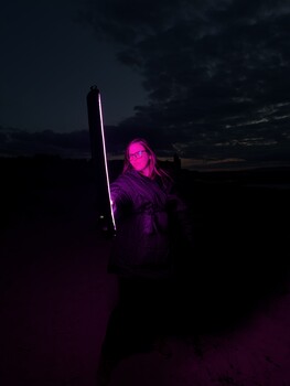 Figure illuminated by neon pink and purple light holding a vertical light saber against twilight sky