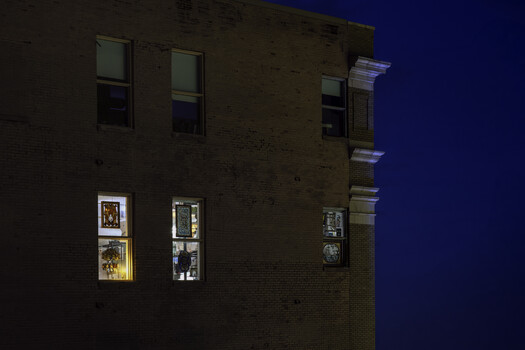 Brick building facade at dusk with illuminated window displays showing framed artwork