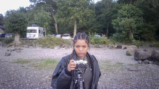 Young woman holding a camera with tripod mount, standing in a gravel camping area surrounded by forest