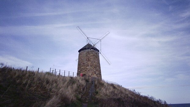 Stone windmill with four sails on a grassy hillside against a cloudy sky