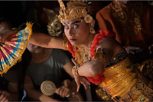 Woman in ornate gold costume and traditional headpiece holds decorative fan during cultural performance