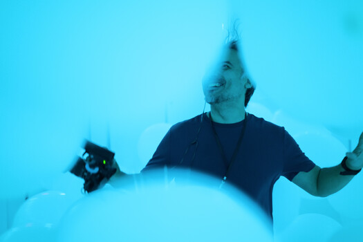 Smiling man in a navy t-shirt looking upward, lit by saturated blue light in a white curved space, holding a black handheld device.