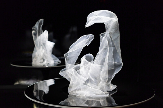 Sheer white fabric sculptures twisted into abstract forms on reflective black pedestals against a dark background.