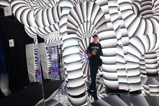 Man holding a camera in an immersive room lined with black-and-white swirling optical illusion columns and a mirrored floor