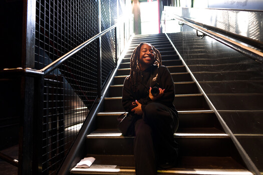 Smiling woman with long locs sitting on a dark metal staircase, dressed in black, eyes closed and laughing.