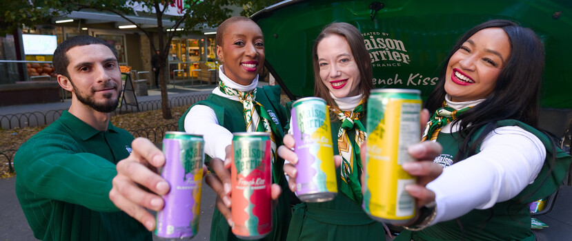 Four people in green uniforms extend colorful canned drinks toward the camera in a toast on a city street.