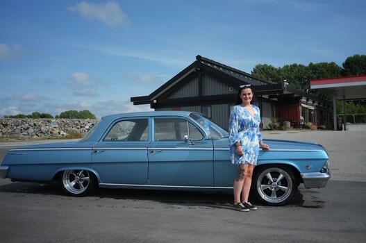 Woman standing beside a restored blue 1950s sedan in an industrial setting