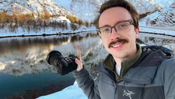 Photographer holding a camera with snow-covered mountains and a frozen river in the background.