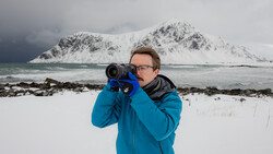 Photographer in blue jacket holding telephoto lens toward snow-covered mountain across water.