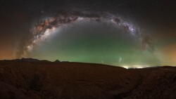 Milky Way arching over a desert landscape with green aurora lights on the horizon.