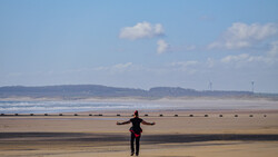 Person with arms outstretched standing alone on a wide, wet sandy beach with gentle waves and distant hills.