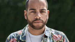 Man with beard wearing floral patterned shirt and white undershirt, photographed outdoors with blurred green background.