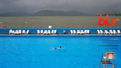 Swimmer in turquoise pool with white loungers and gray sea beyond. Mountains visible across the water.