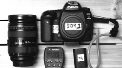 Canon EOS 5D Mark IV camera body with Sigma 24-70mm lens, Profoto flash trigger, and accessories on white wood surface.