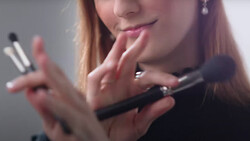 Woman with reddish-brown hair holding makeup brushes and examining cosmetic products near her face.