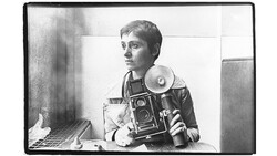 Black and white self-portrait of a photographer holding a large format camera and light meter.