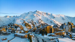 Snow-covered alpine resort nestled below a dramatic snow-capped mountain peak under clear blue sky.