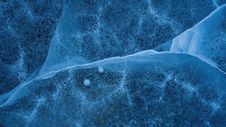 Aerial view of a frozen lake with thin cracks in the ice and scattered snow across the surface.