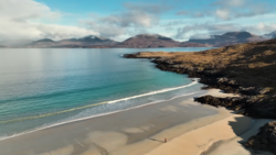 Coastal beach with turquoise water, golden sand, and dark rocky outcrops under blue sky with mountains in distance.