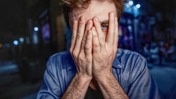Man with reddish-brown hair covering his face with both hands, wearing a blue shirt against a blurred urban night background.