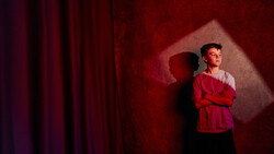 Young man in red shirt standing with arms crossed against red wall with dramatic shadow and stage lighting.