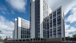 Modern waterfront residential buildings with mixed-height towers and wooden base structure under blue sky with clouds.